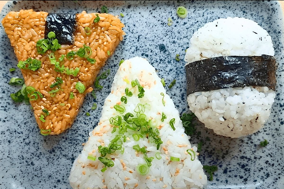 Three onigiri on a speckled blue tray: soy glazed, plain with spring onion, and one wrapped in nori