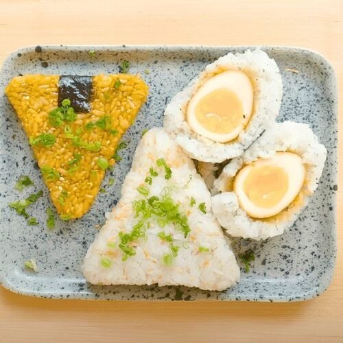 Overhead view of three onigiri on a speckled blue tray: soy glazed, plain with spring onion, and a soy egg wrapped in rice