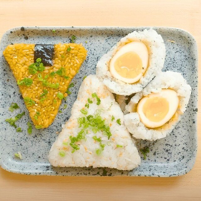 Overhead view of three onigiri on a speckled blue tray: soy glazed, plain with spring onion, and a soy egg wrapped in rice