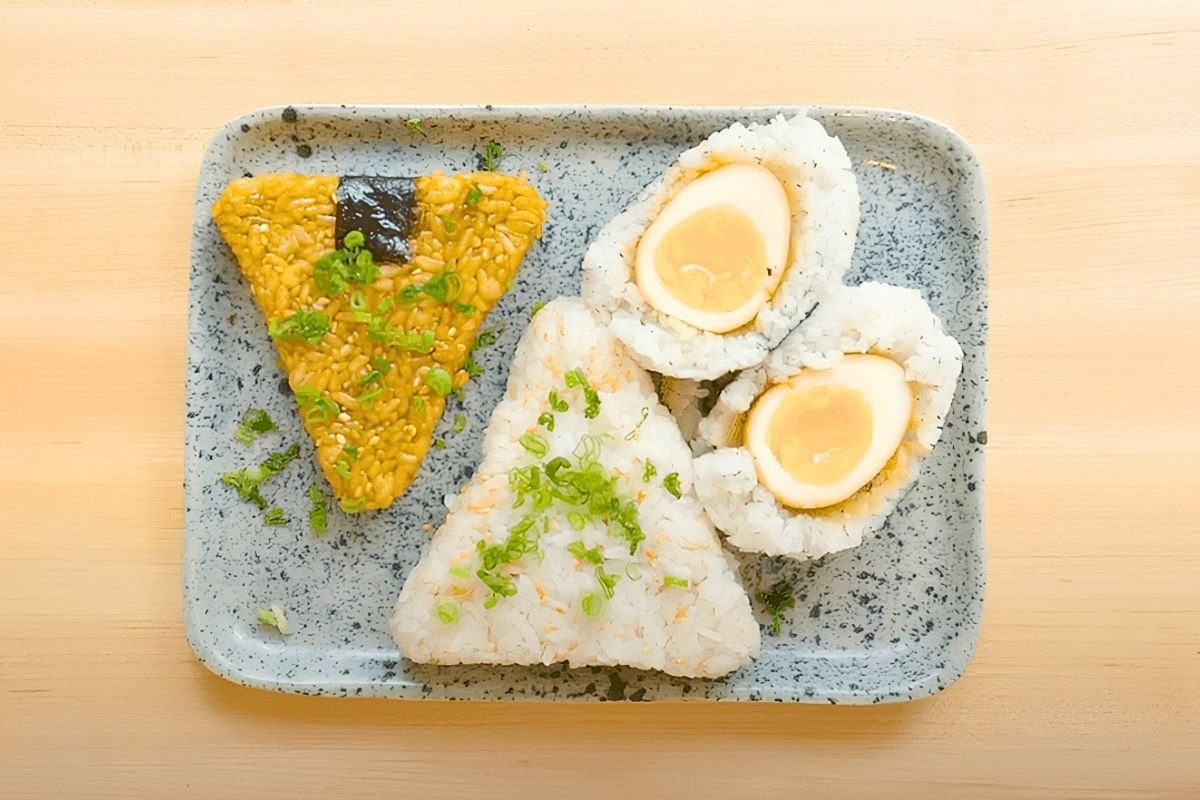 Overhead view of three onigiri on a speckled blue tray: soy glazed, plain with spring onion, and a soy egg wrapped in rice