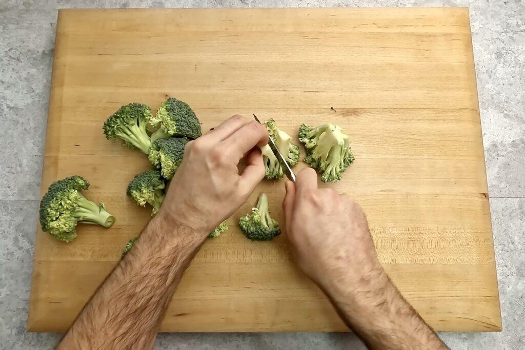 Hands pulling broccoli florets apart on a wooden chopping board with small florets scattered around