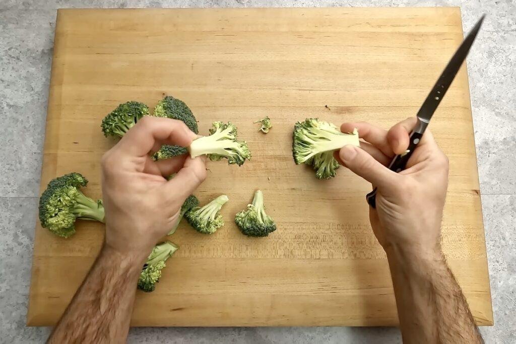 Hands cutting broccoli into florets with a knife on a wooden chopping board