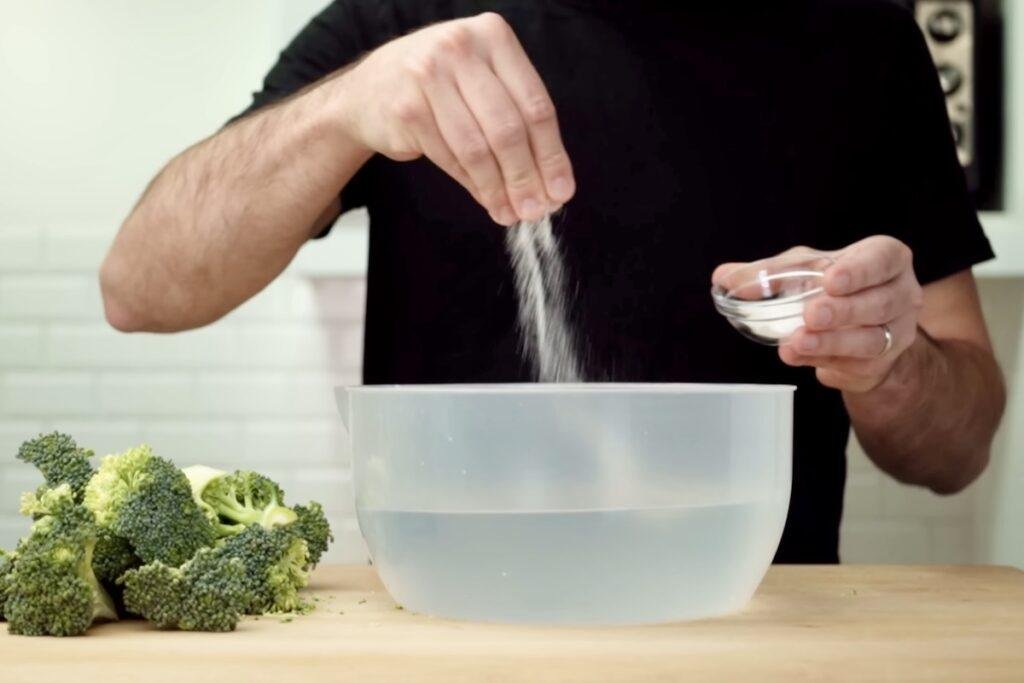 Pouring salt into a bowl of cold water with a head of broccoli beside it