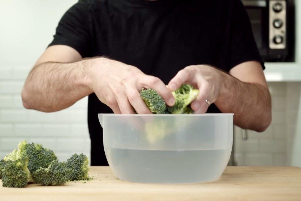 Hands lowering broccoli florets into a bowl of salted water