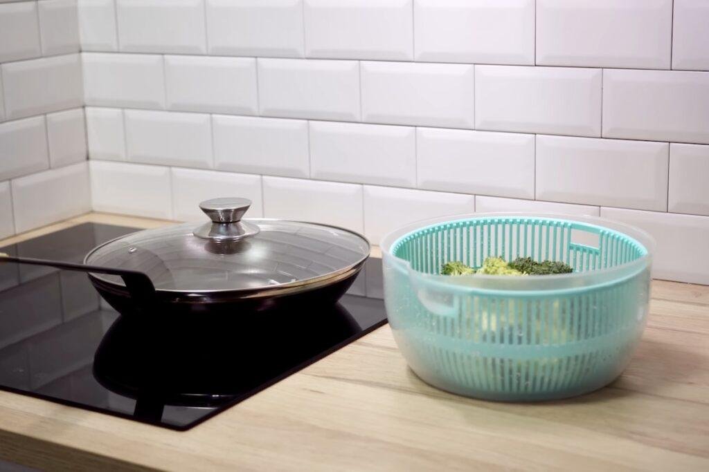 Teal salad spinner with broccoli inside next to a lidded pan on an induction hob