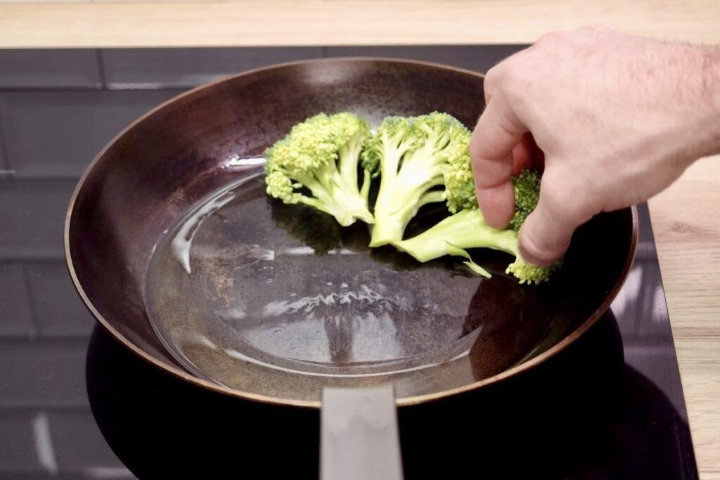 Hand placing a broccoli floret cut side down in a frying pan of hot oil