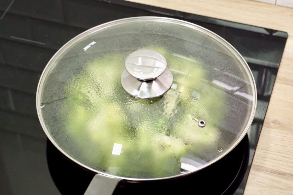 Glass lid covering a pan of broccoli on an induction hob with steam misting the glass
