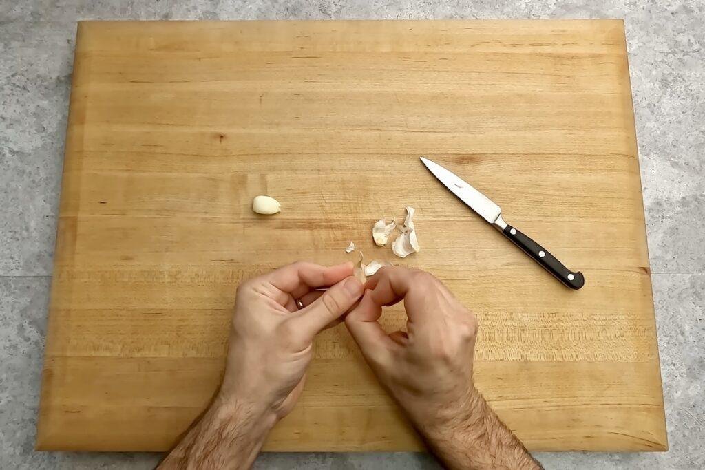 Hands peeling garlic cloves on a wooden chopping board next to a knife