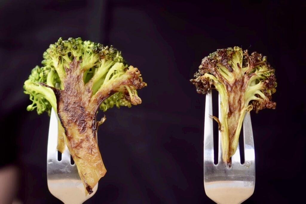 Two broccoli florets on forks side by side, the left charred and bright green, the right darker and drier