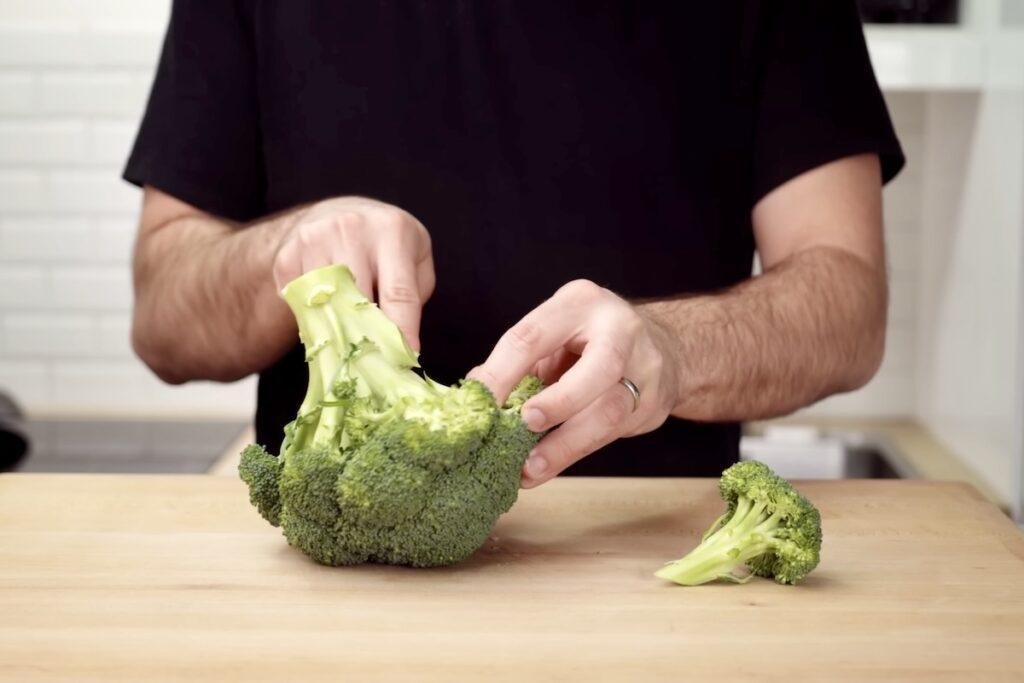 Hands holding a whole head of broccoli on a chopping board with a cut floret beside it