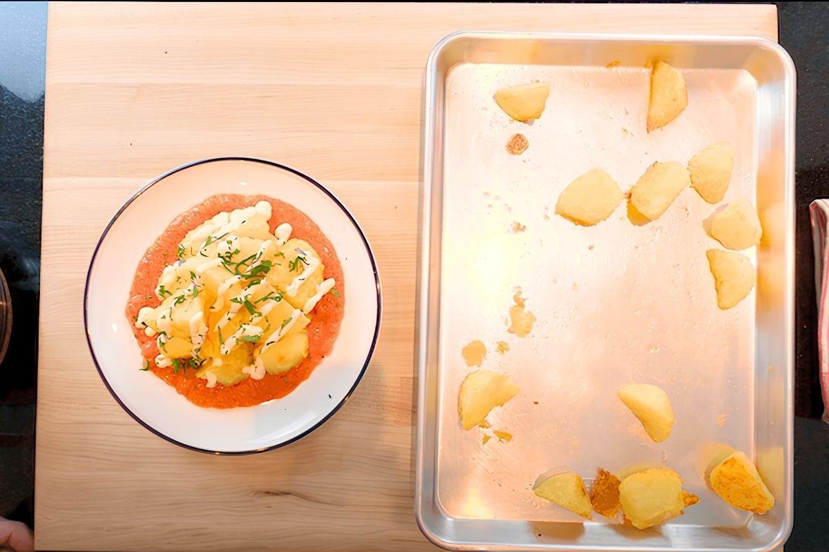Plate of patatas bravas beside a tray of crispy potato pieces on a wooden board