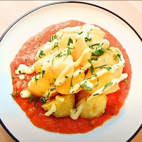 Overhead view of patatas bravas on a white plate with tomato sauce, aioli and parsley