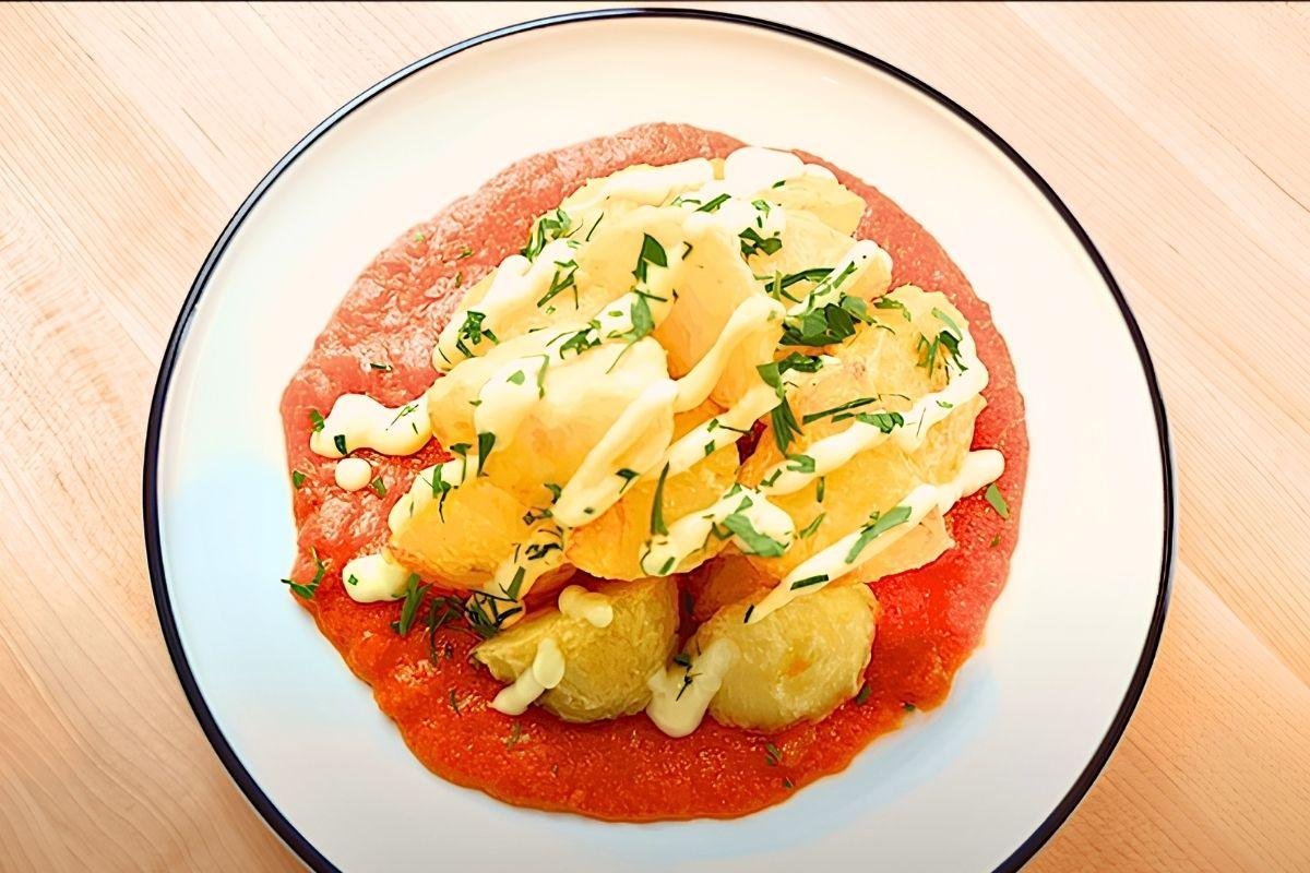Overhead view of patatas bravas on a white plate with tomato sauce, aioli and parsley