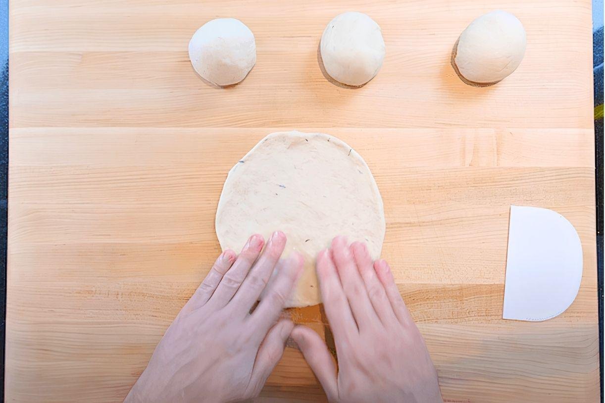 Hands flattening a ball of pitta dough on a wooden board beside three more dough balls