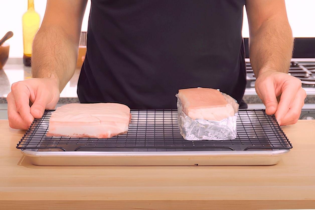 Two pieces of pork belly, one wrapped in foil, resting on a wire rack over a tray