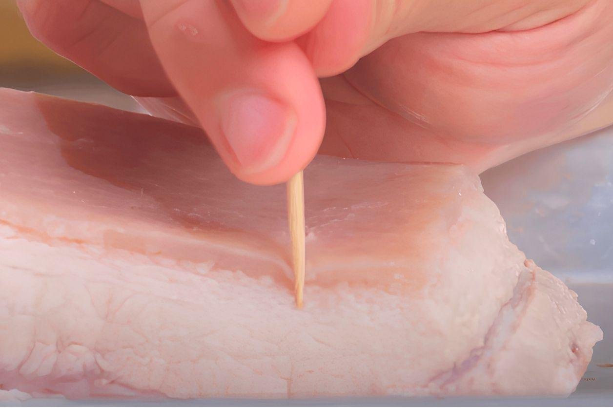 Hand piercing the skin of raw pork belly with a toothpick