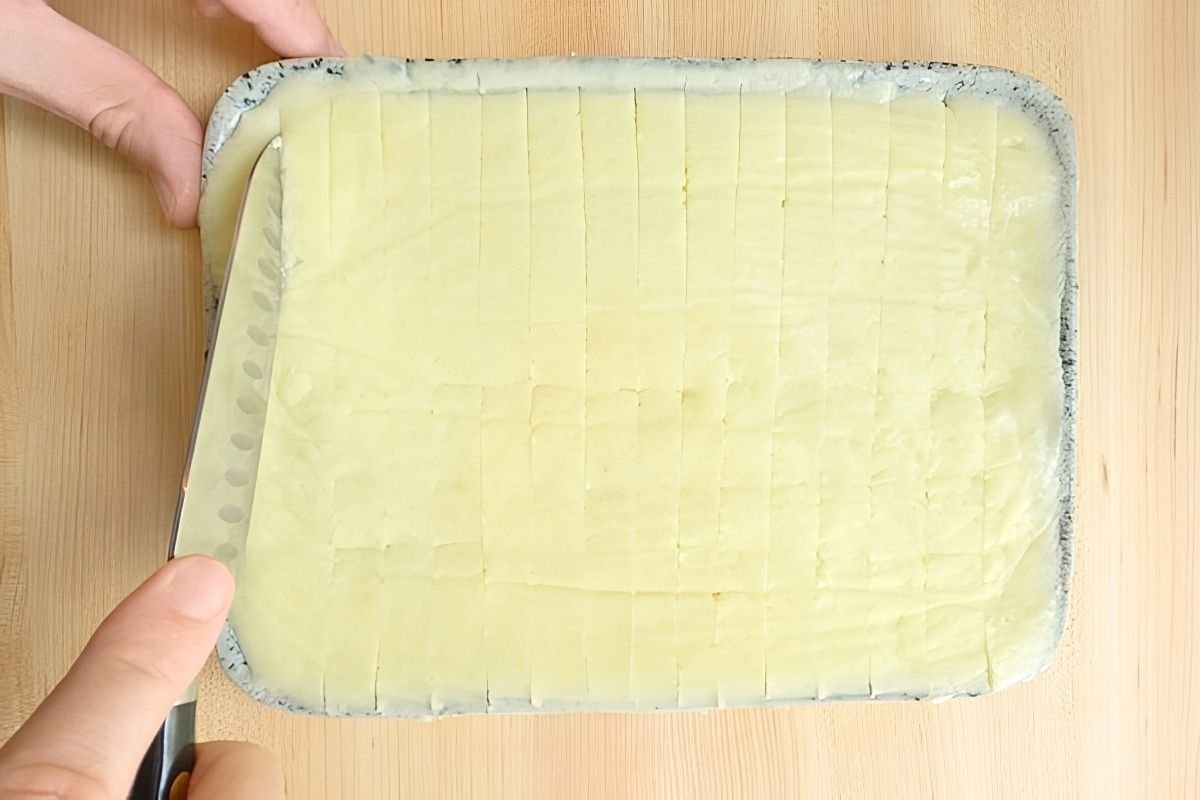 A hand holds a knife above a tray of cut, creamy potatoes, preparing them for the ultimate fries recipe.
