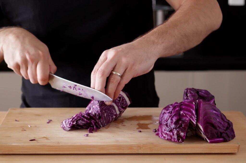 Person slicing red cabbage with a knife on a wooden board, with cabbage quarters beside