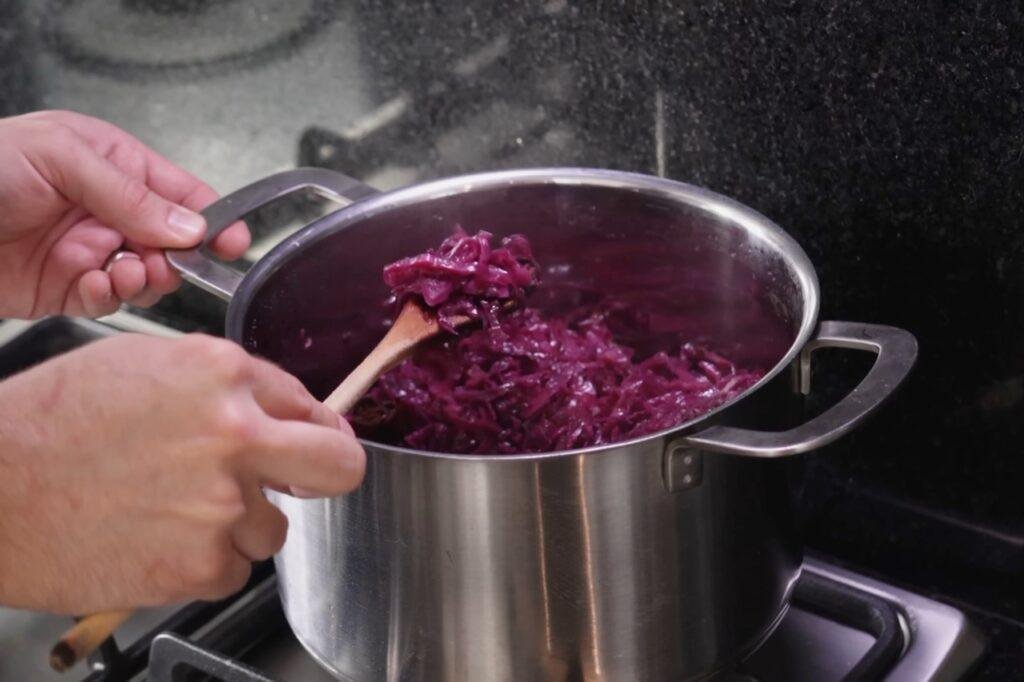 Hand stirring shredded red cabbage in a steel pot with a wooden spoon
