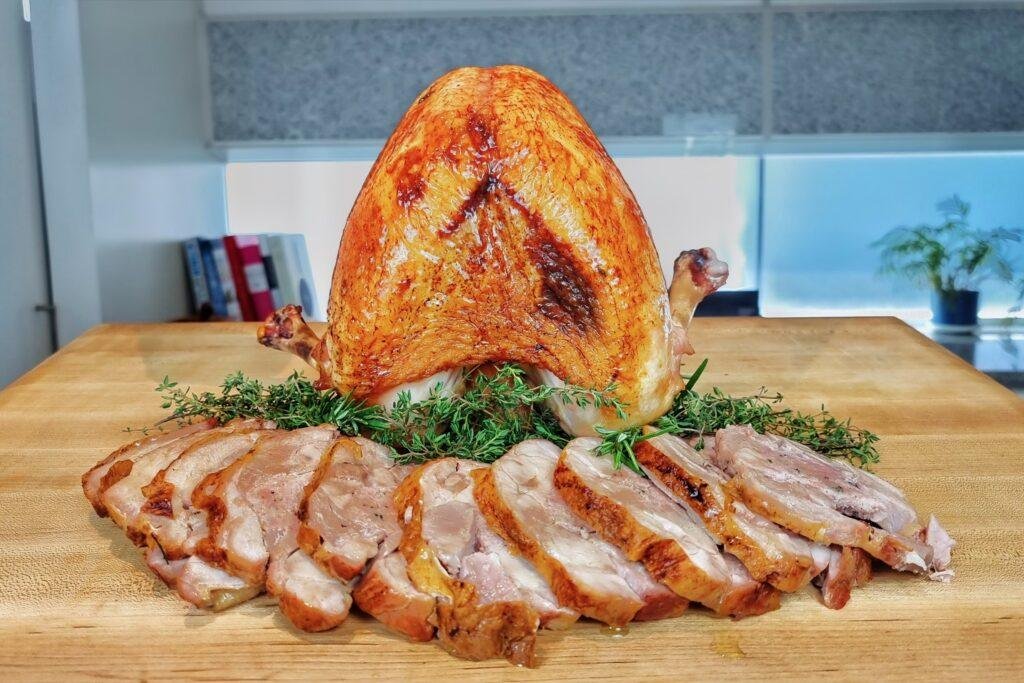 Roast turkey breast with golden skin above sliced turkey on a bed of rosemary on a wooden board