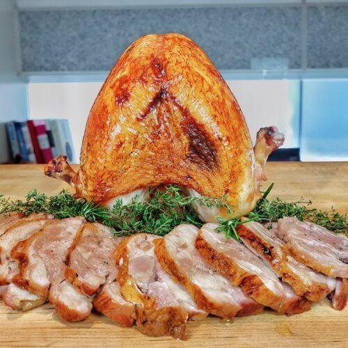 Roast turkey breast with golden skin above sliced turkey on a bed of rosemary on a wooden board