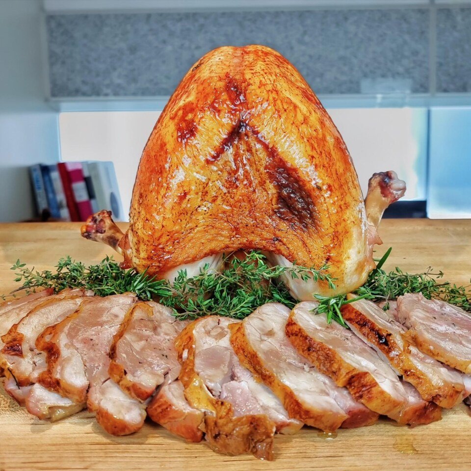 Roast turkey breast with golden skin above sliced turkey on a bed of rosemary on a wooden board
