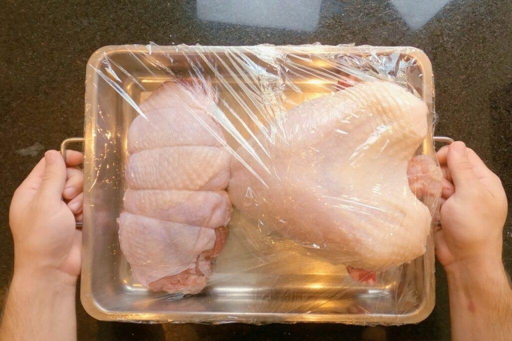 Overhead view of hands holding a roasting tin of raw turkey pieces wrapped in cling film