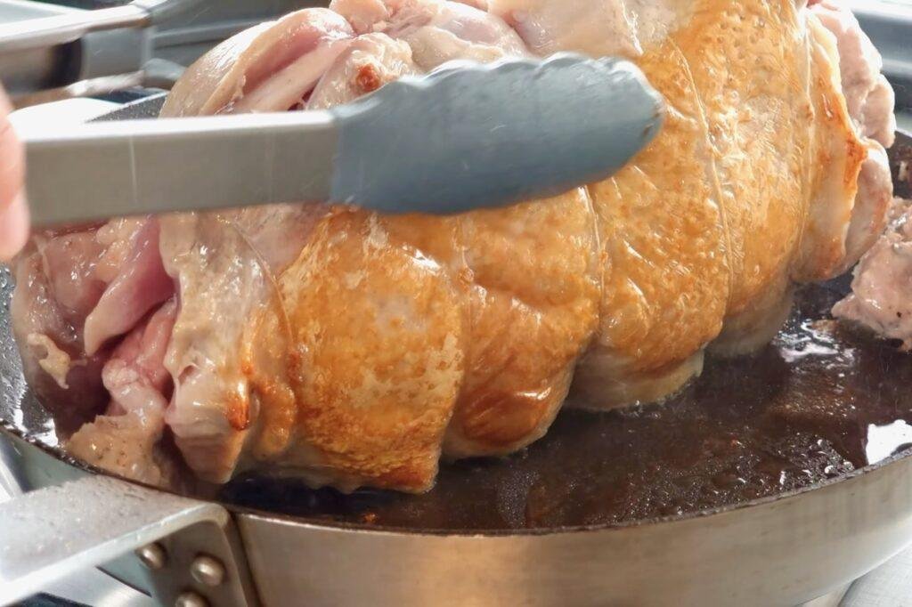 Tongs turning a golden roasted stuffed turkey leg in a frying pan