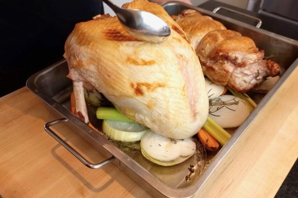 Spoon basting a golden roast turkey crown in a roasting tin with vegetables