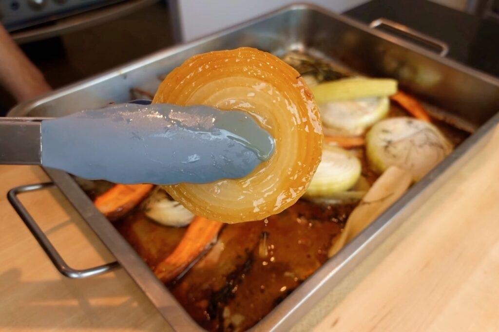 Tongs lifting a roasted onion slice from a roasting tin of vegetables and pan juices