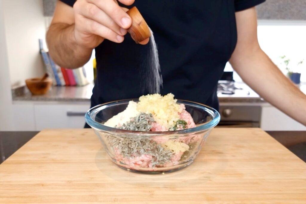 Person sprinkling seasoning into a glass bowl of pink meat, herbs and a pale mixture