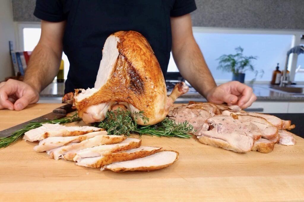 Person carving a golden roast turkey crown, with slices laid on rosemary on a wooden board