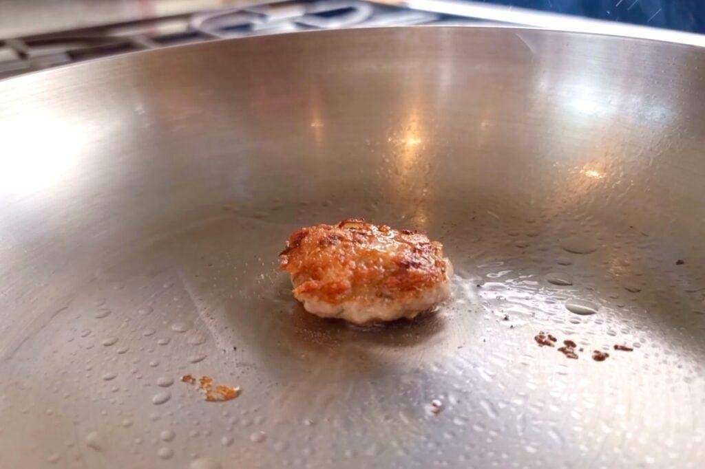 Small browned patty frying in a large steel pan