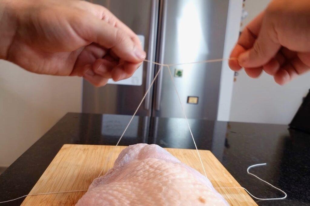 Hands pulling string tight around a raw turkey joint on a wooden board