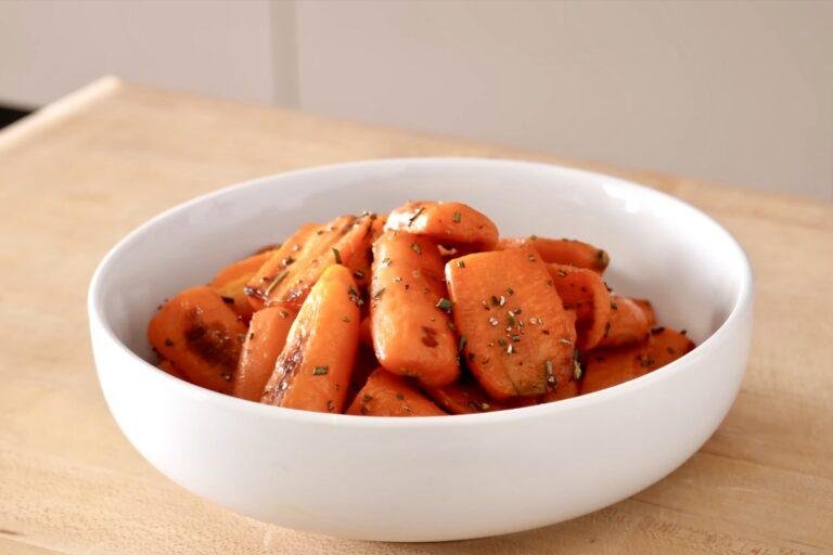 White bowl of glazed roasted carrot pieces with herbs on a wooden counter