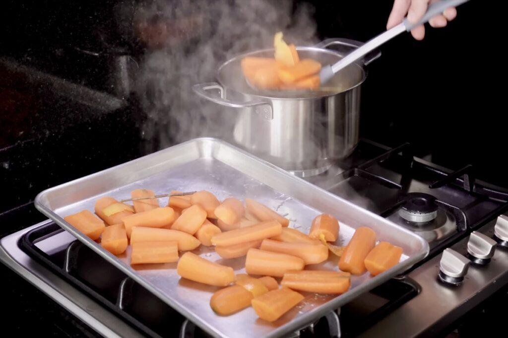 Par-boiled carrot pieces lifted from a steaming pot onto a metal baking tray on the hob