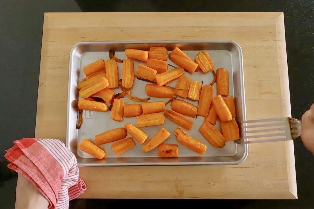 Overhead view of cut carrot pieces roasted on a metal baking sheet on a wooden board, with a spatula at the right and a striped towel at the left