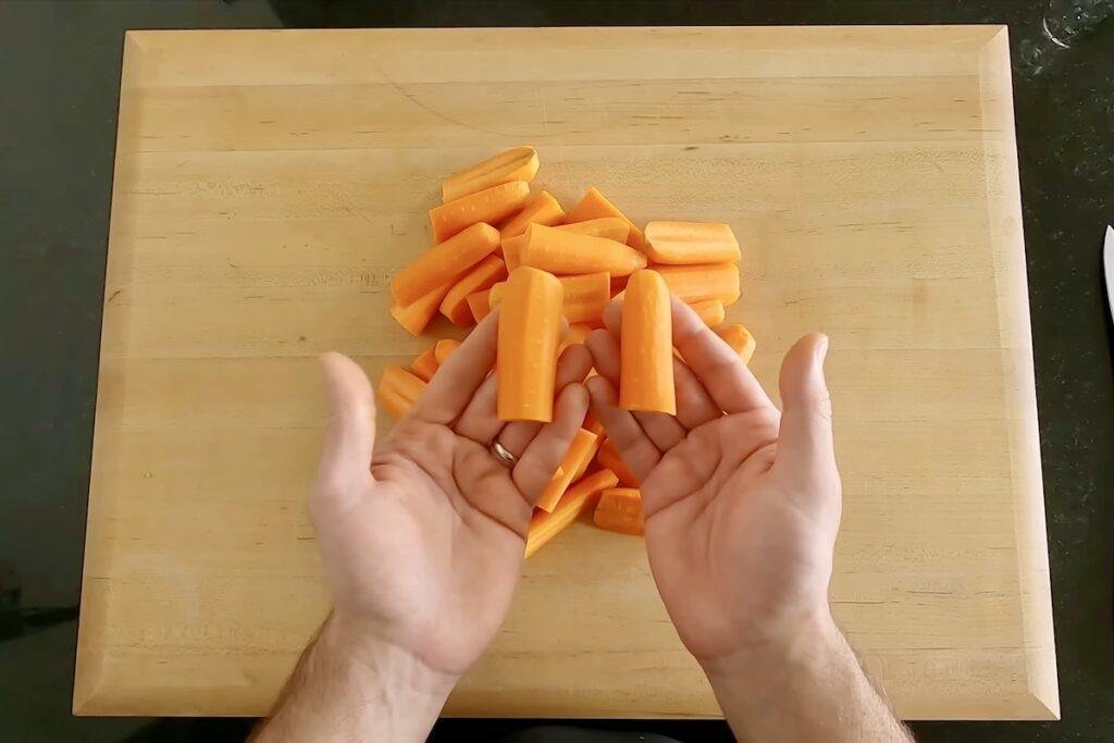 Two hands held open over a wooden cutting board with a pile of carrots cut into short sticks