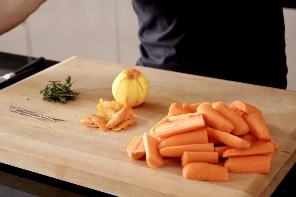 Cutting board with sliced carrots, a sprig of rosemary and a whole lemon next to a pile of lemon peel