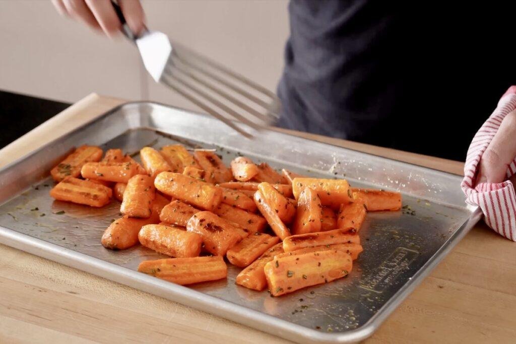 Browned roasted carrots on a baking sheet being lifted with a spatula, with a striped oven towel at the right