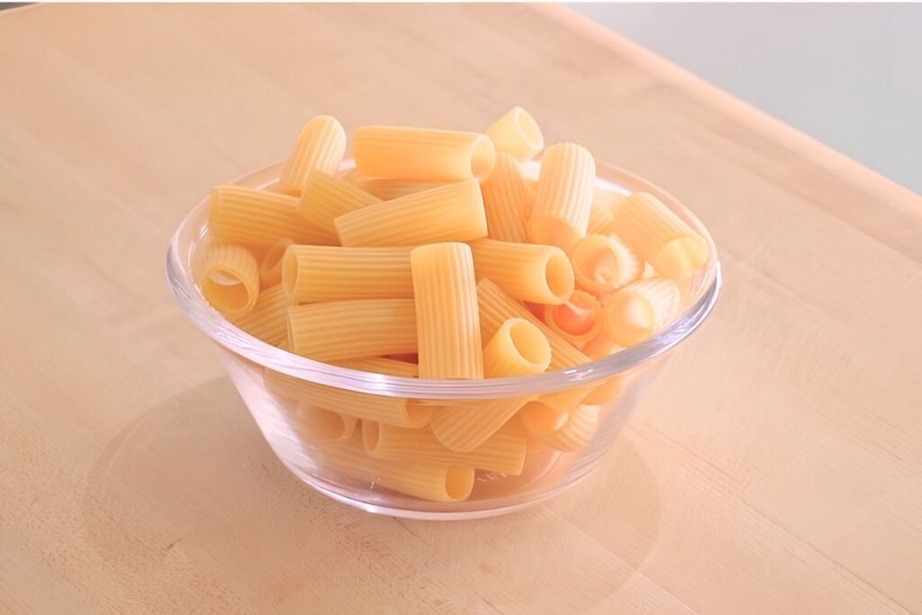 Glass bowl of dry rigatoni pasta on a wooden board