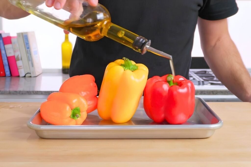Olive oil being poured over red, yellow and orange peppers on a baking tray