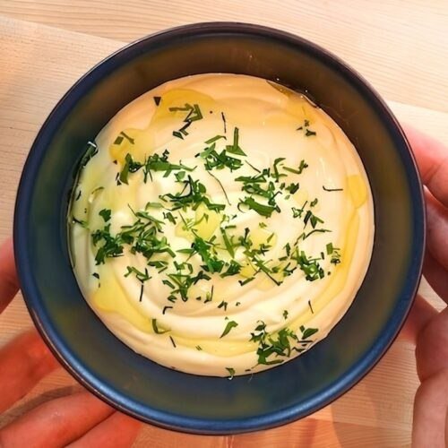 Creamy aioli topped with olive oil and fresh herbs, served in a blue bowl on a wooden surface.