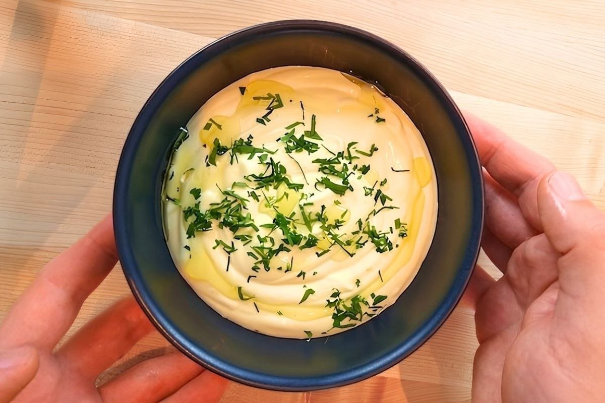 Creamy aioli topped with olive oil and fresh herbs, served in a blue bowl on a wooden surface.