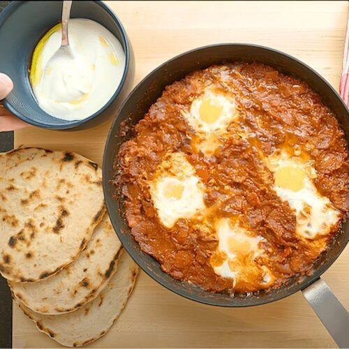Shakshuka in a pan with flatbreads and a small bowl of yoghurt