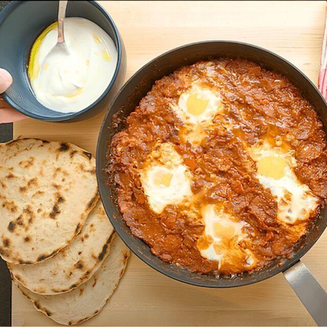 Shakshuka in a pan with flatbreads and a small bowl of yoghurt