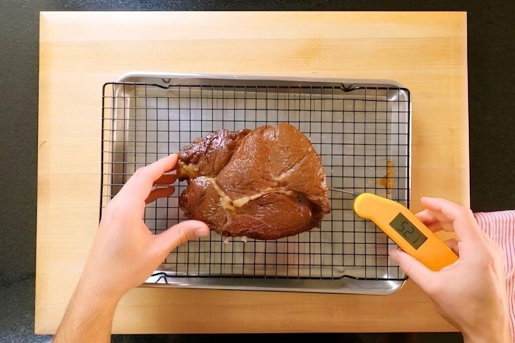 Hand holding a thermometer next to a seared steak on a wire rack over a tray