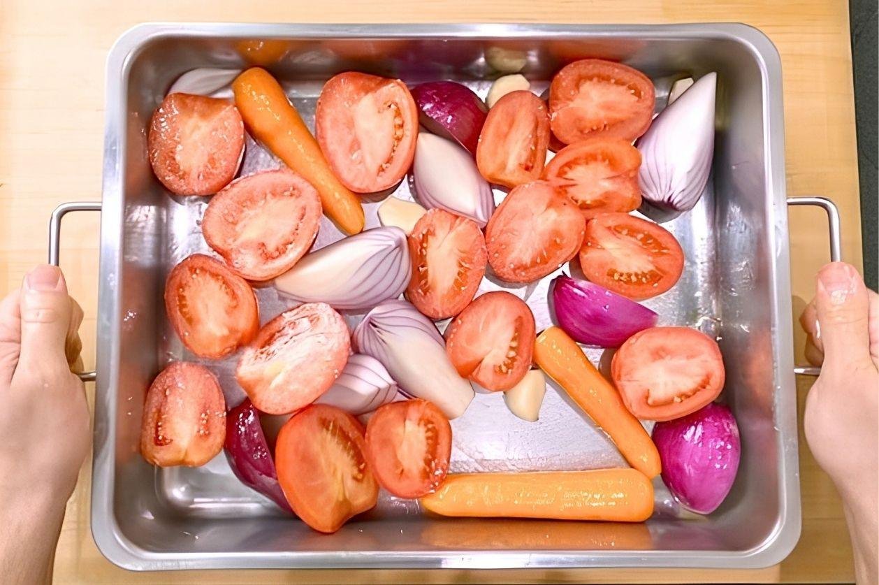 Overhead view of a roasting tin of halved tomatoes, red onions and carrots ready to roast
