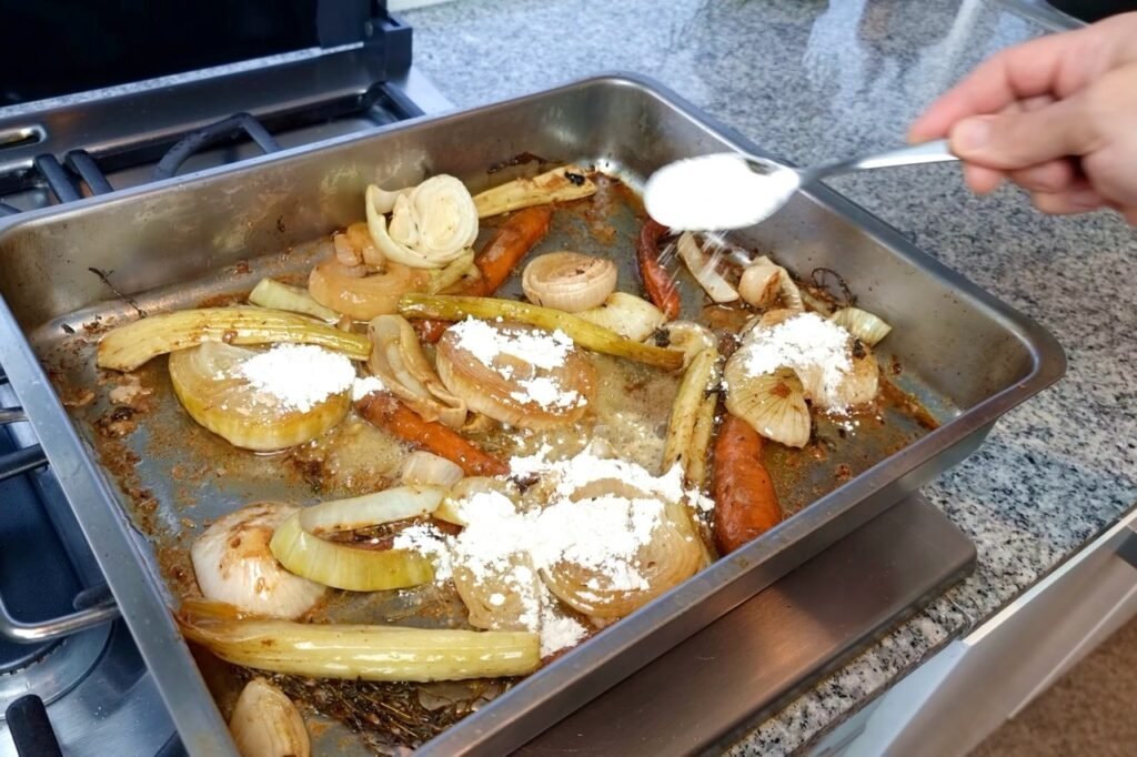 Spoon sprinkling flour over roasted vegetables in a roasting tin