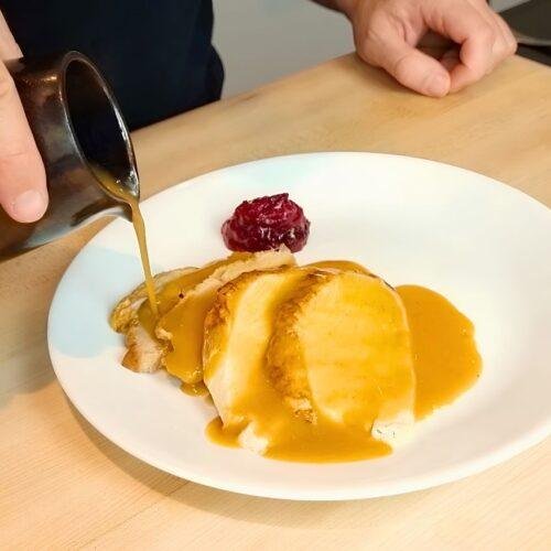 Gravy being poured from a jug over sliced turkey on a white plate with cranberry sauce